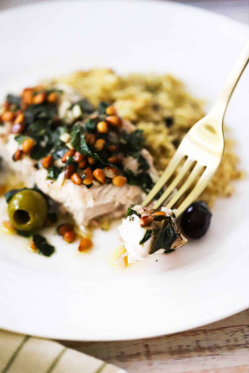 A gold fork holding up a piece of baked red snapper taken from a plate holding the fillet of fish covered in olives, pine nuts, and garlic. 