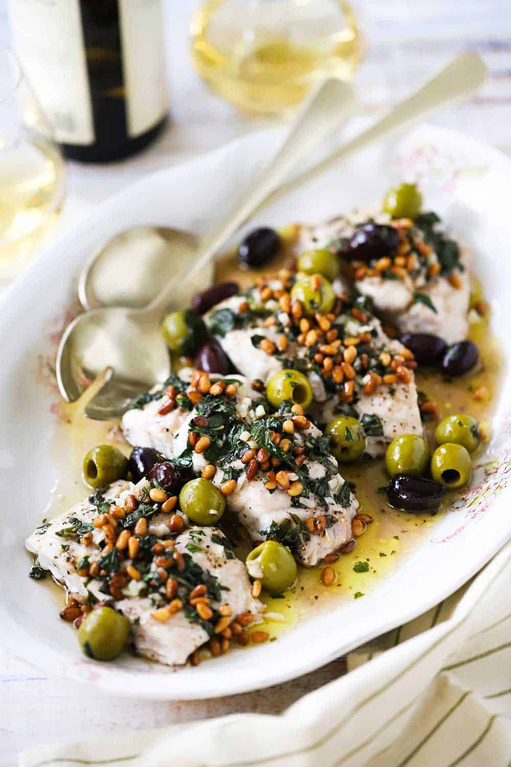 An oval white platter filled with several filets of Mediterranean-style baked red snapper all next to a glass and bottle of white wine. 