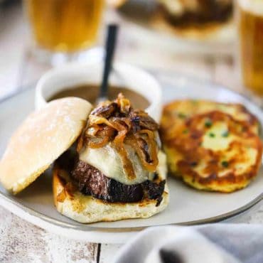 A white plate filled with a pot roast slider topped with caramelized onions sitting next to two potato cakes, with a small bowl of gravy next to it, as well.