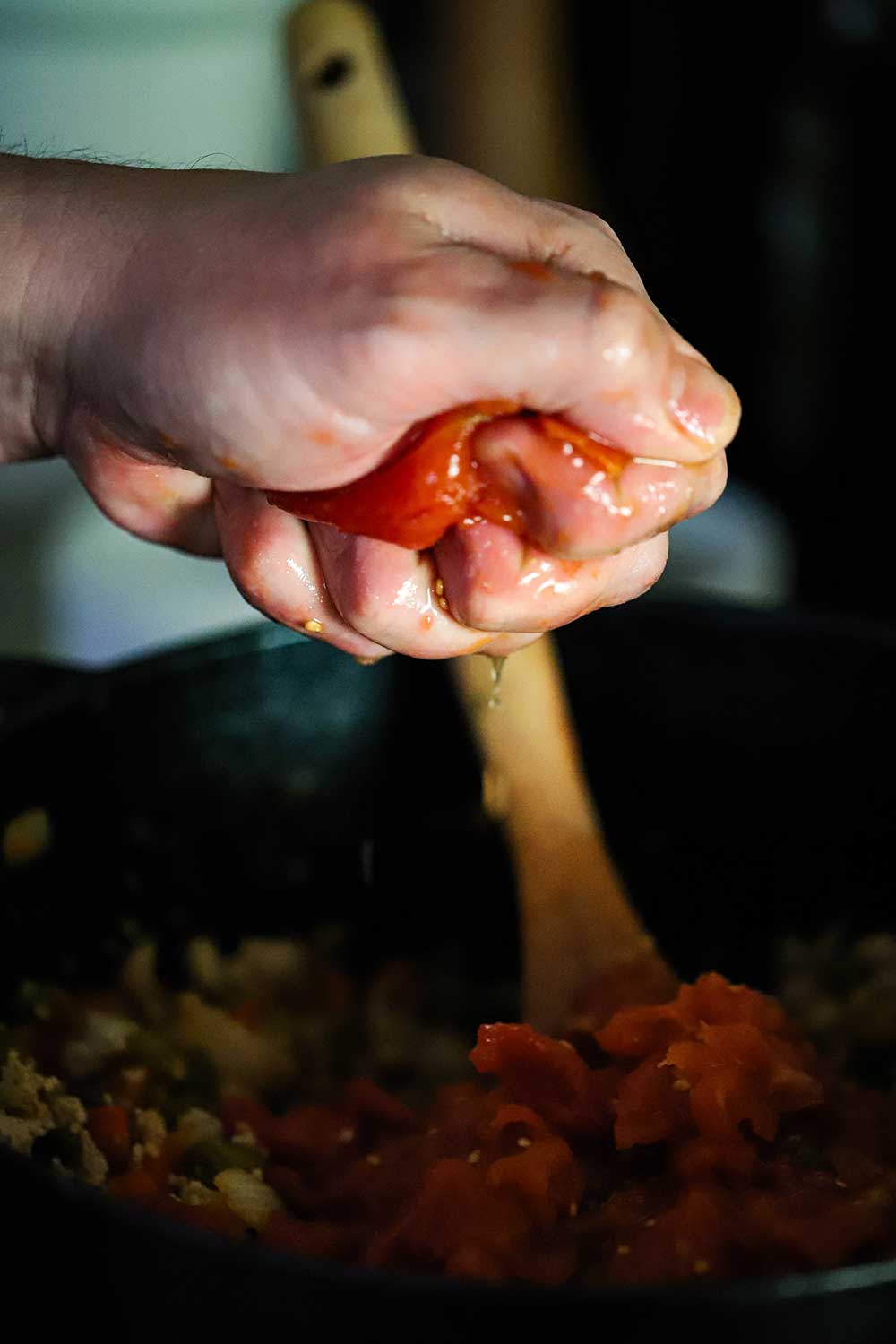 A hand squeezing a whole tomato over a cast-iron pot full of cooked ground turkey and vegetables.
