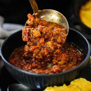 A helping of turkey chili being ladled into a dark blue bowl next to a piece of cornbread.