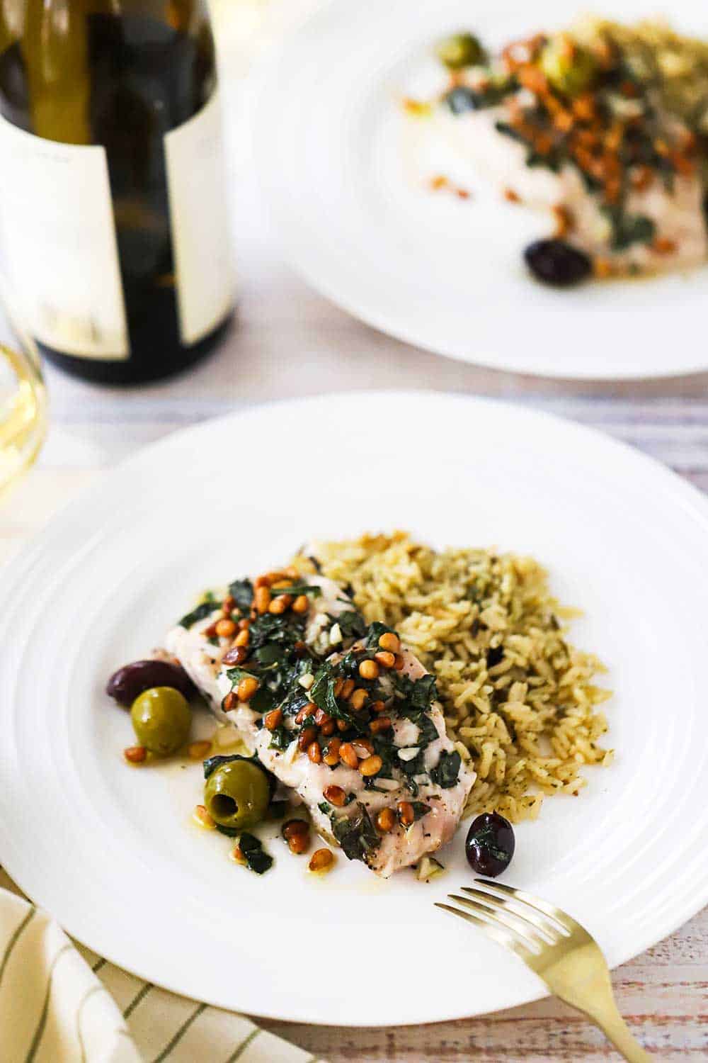 Two white dinner plates filled with Mediterranean-style baked red snapper and a mound of cooked wild rice, both sitting next to a bottle of wine. 