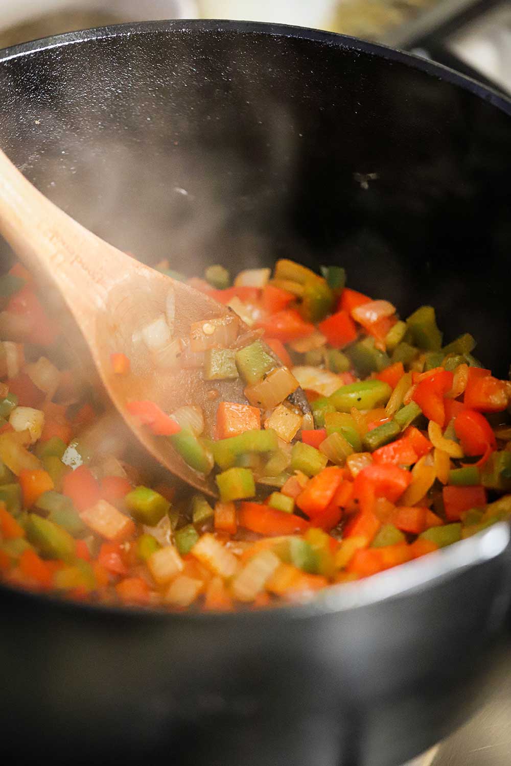 A large black cast-iron pot filled with chopped green and red peppers simmering with a large wooden spoon stirring it all.