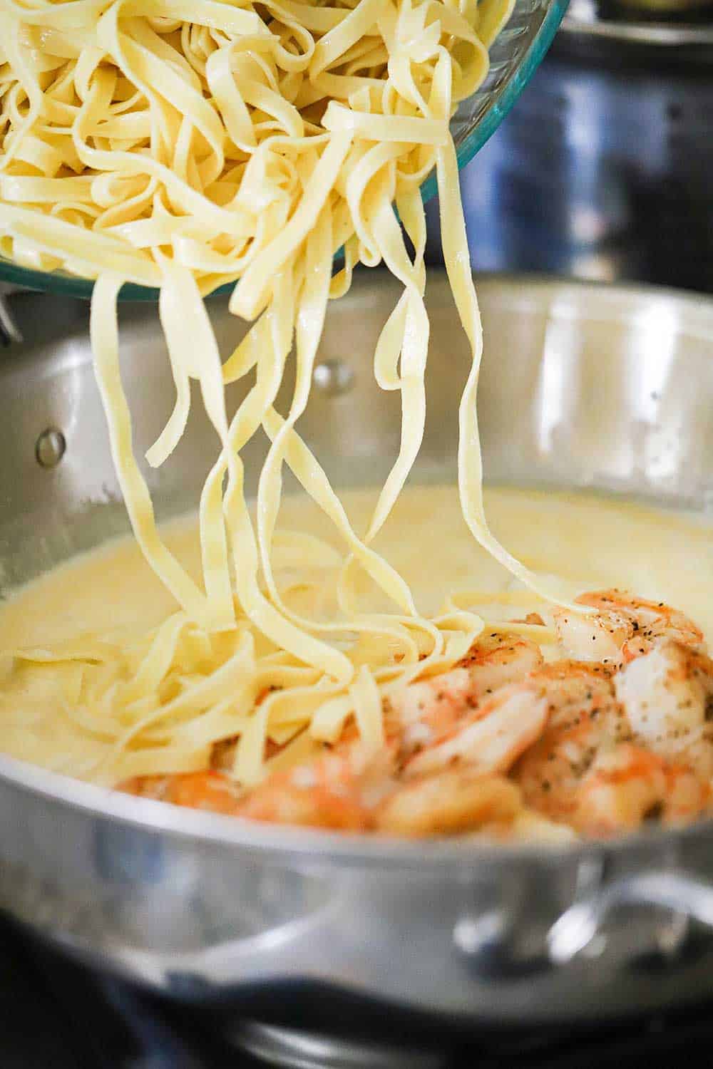 A glass bowl filled with cooked fettuccine is being transferred into a skillet filled with a cream sauce and sautéd shrimp. 