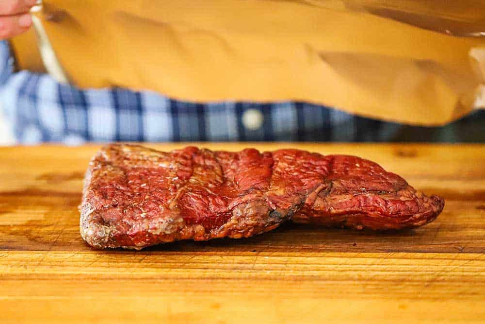 A fully cooked cut of Wagyu chuck short ribs on a cutting board with a piece of foil being held over it ready to be lowered onto the meat. 