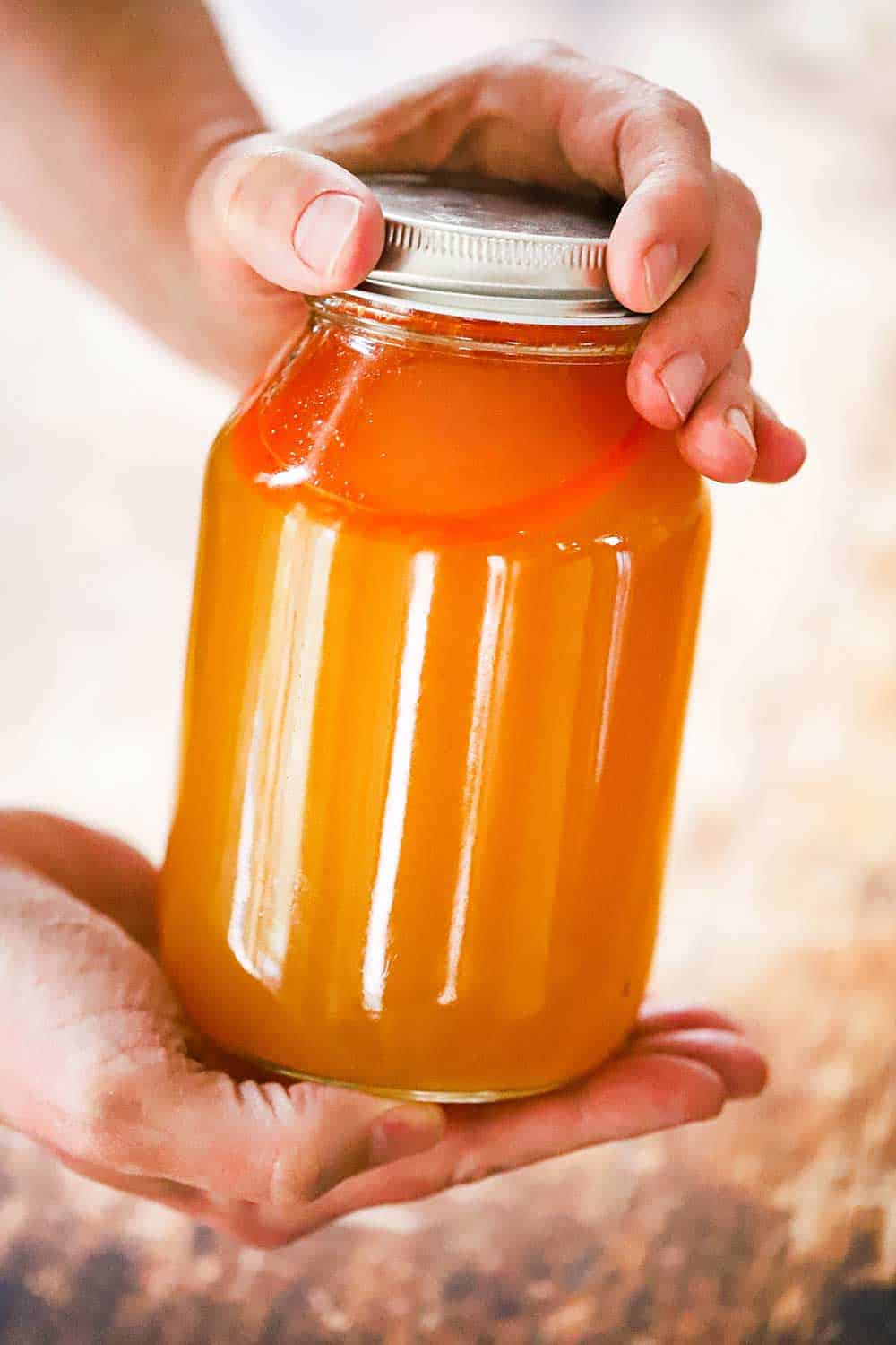 Two hands holding a glass jar with a lid filled with shellfish stock.