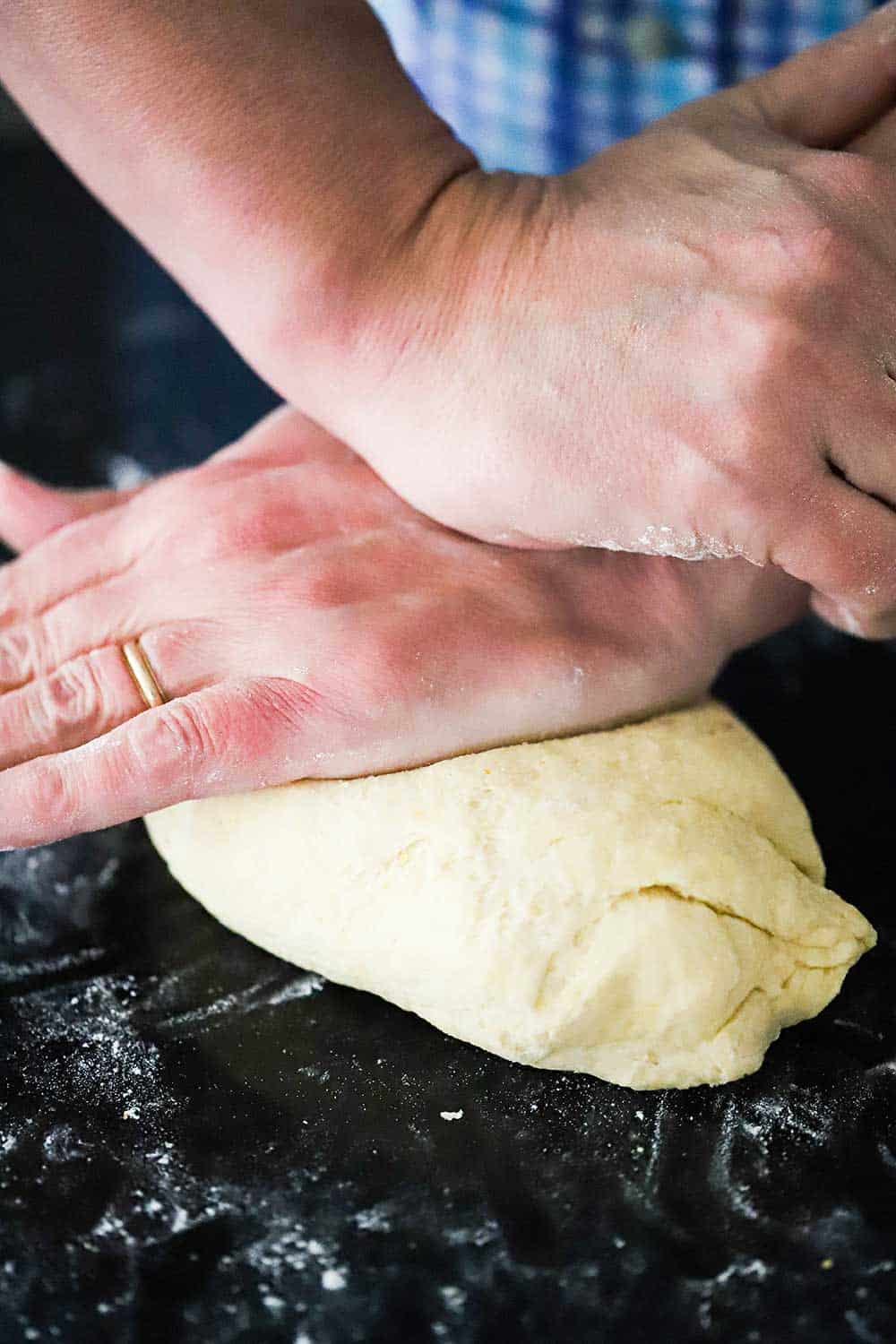 Two hands, one pressing on top of the other, are kneading pizza dough on a black lightly floured marble surface. 