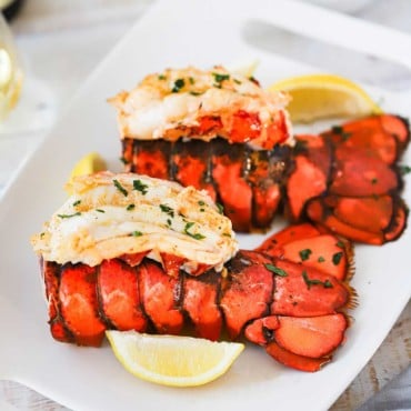 Two fully cooked lobster tails with meat on top of the lobster tail, all sitting on a platter next to lemon wedges and melted butter.
