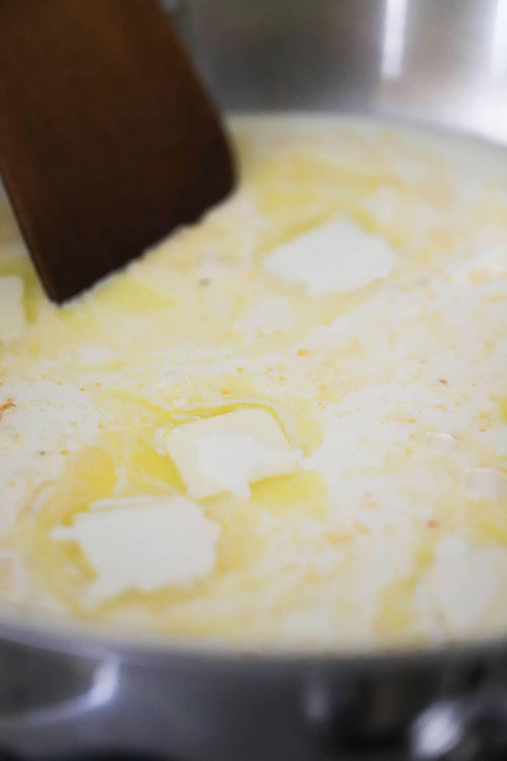 A large stainless steel skillet filled with cream and small pallets of butter that are melting and being swirled by a wooden spatula. 