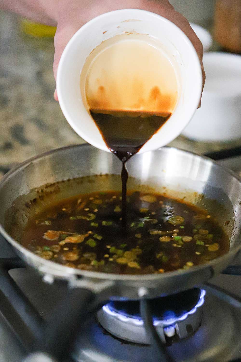 A person pouring a soy sauce and cornstarch mixture from a small bowl into a small skillet with a simmering dark-colored sauce in it. 