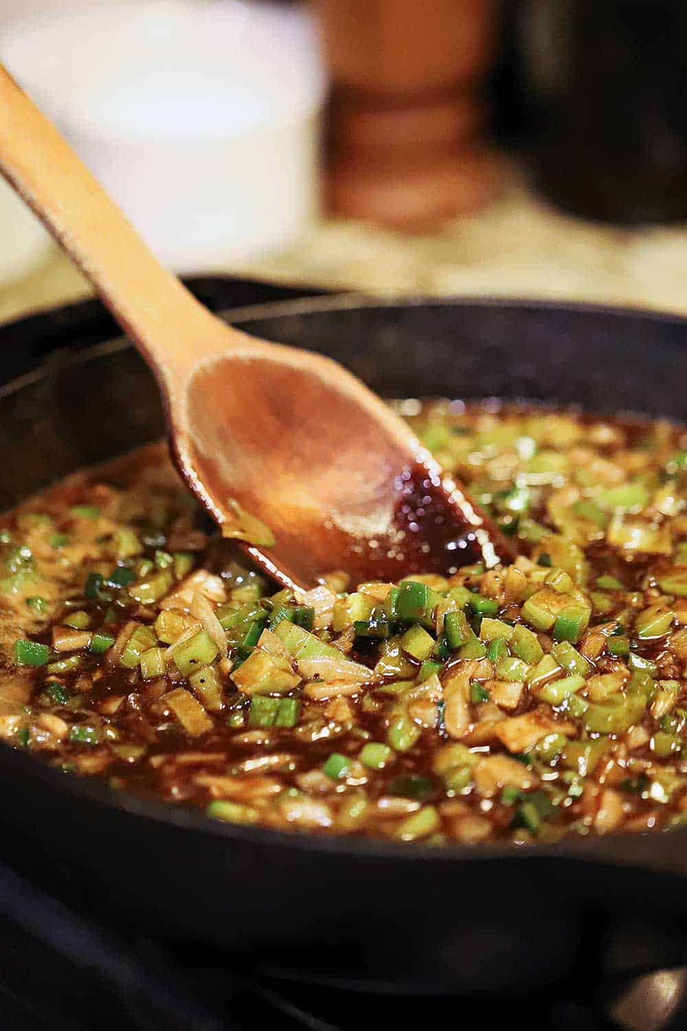 A large black cast -iron skillet filled with a dark roux and chopped vegetables being stirred by a wooden spoon. 