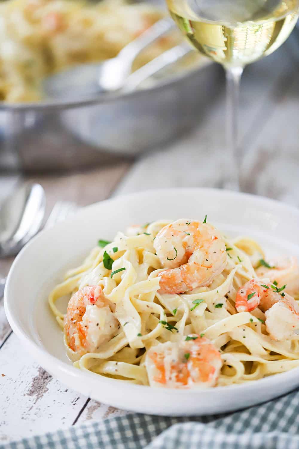 A white individual pasta bowl filled with shrimp fettuccine alfredo next to a large silver skillet filled with the same. 