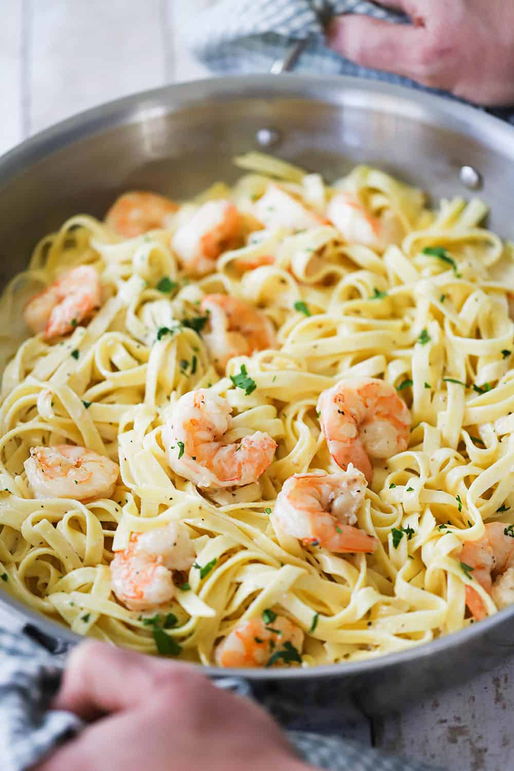 Two hands holding a large stainless steel skillet filled with shrimp fettuccine alfredo. 