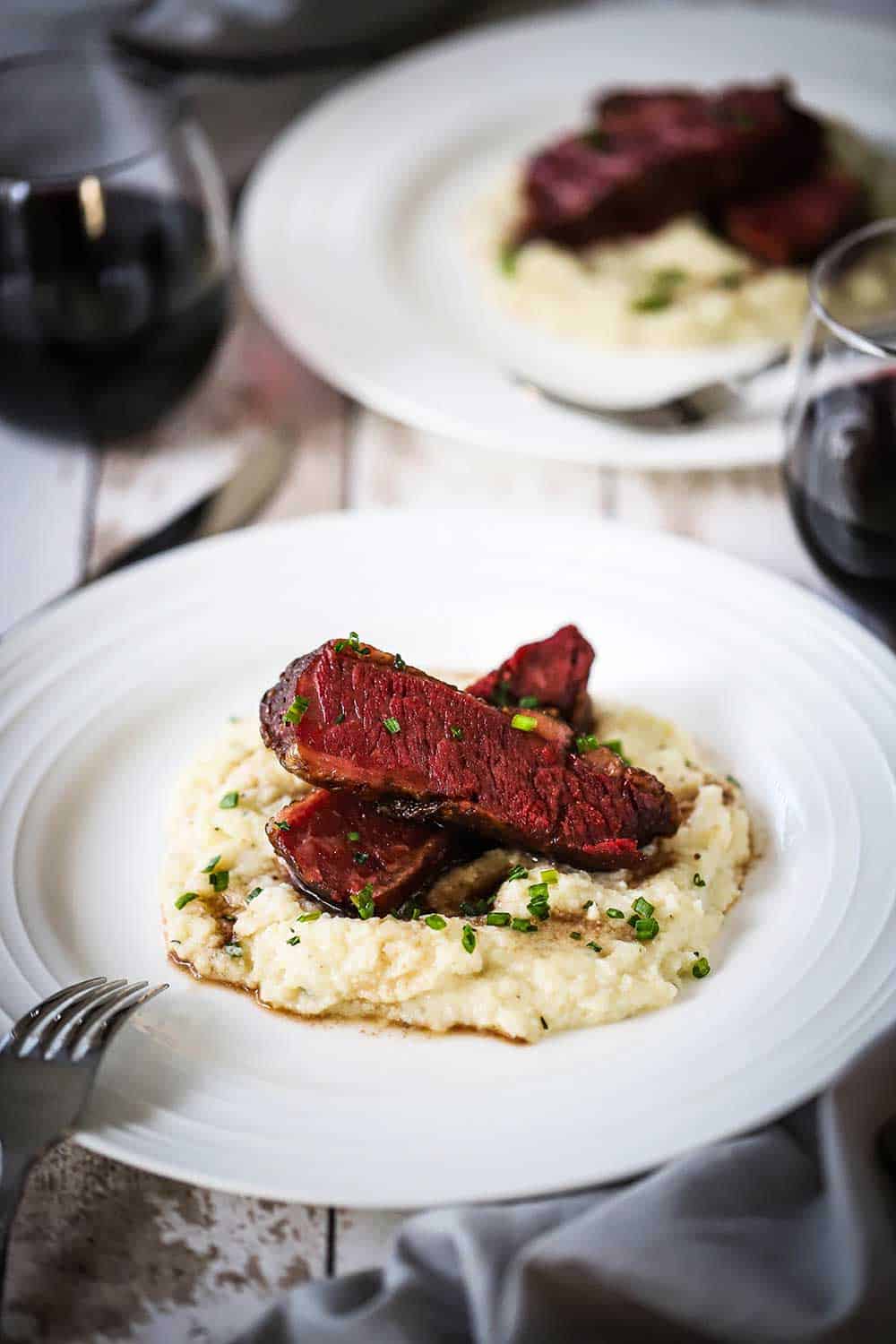 Two white dinner plates, side-by-side, both filled with a bed of mashed cauliflower, topped with two strips of Wagyu chuck short ribs. 