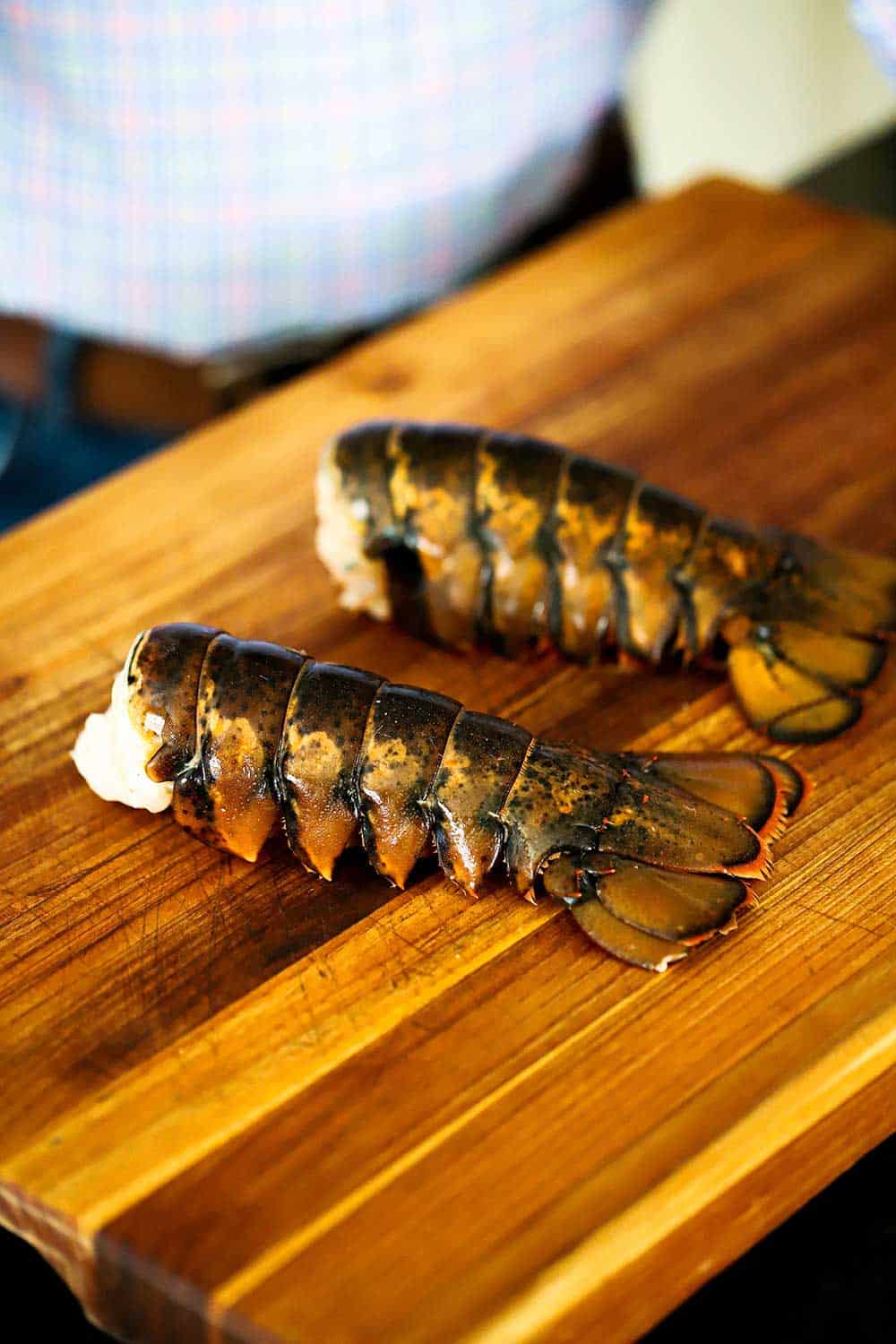 Two half-pound uncooked lobster tails sitting on a large wooden cutting board. 