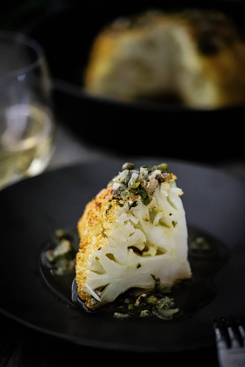 A slice of roasted whole cauliflower sitting on a black plate and topped with a wine butter herb sauce.