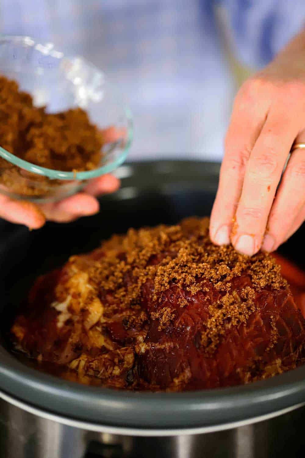 A person using their fingers to sprinkle dark brown sugar all over the top of a spiral ham that has been placed inside a slow-cooker. 