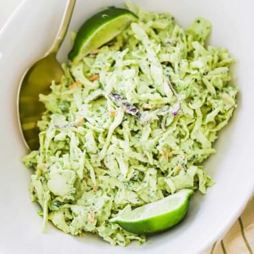 An oval white serving bowl filled with avocado-lime slaw with a gold spoon inserted on the side and a lime wedge on the other side.