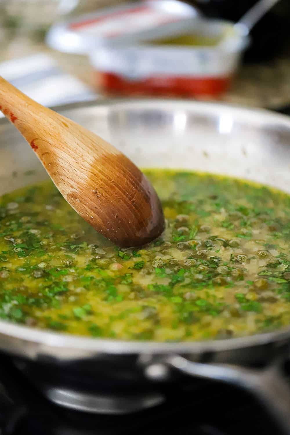 A large stainless steel skilled filled with a butter wine and herb sauce being stirred by a wooden spoon.