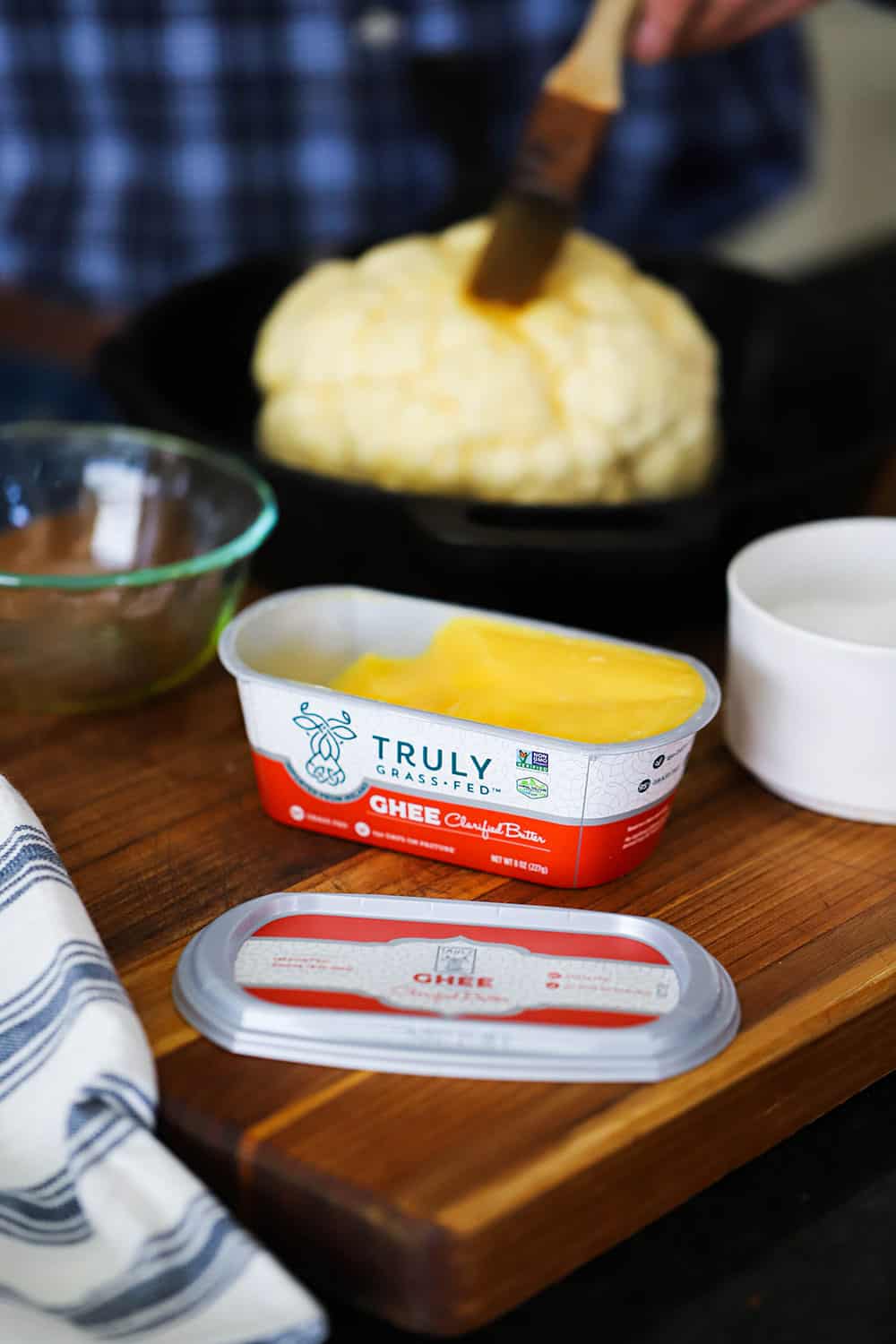 An opened tub of ghee clarified butter in front of a cast-iron skillet filled with a head of cauliflower being buttered.