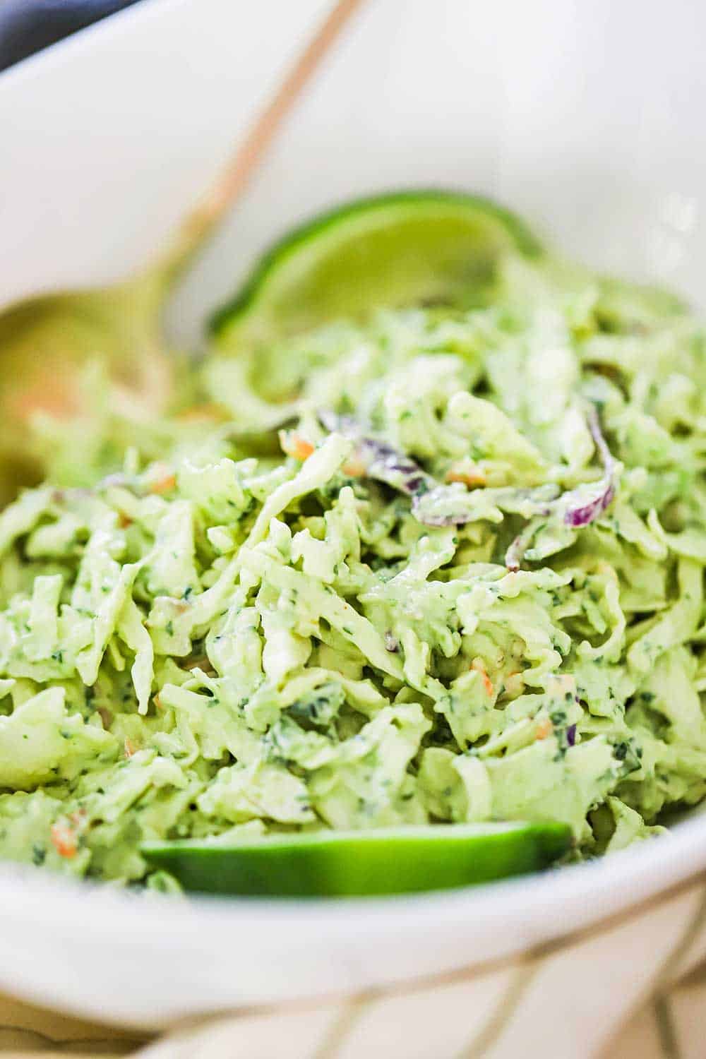 An oval white serving bowl filled with avocado-lime slaw with a gold spoon inserted on the side and a lime wedge on the other side. 