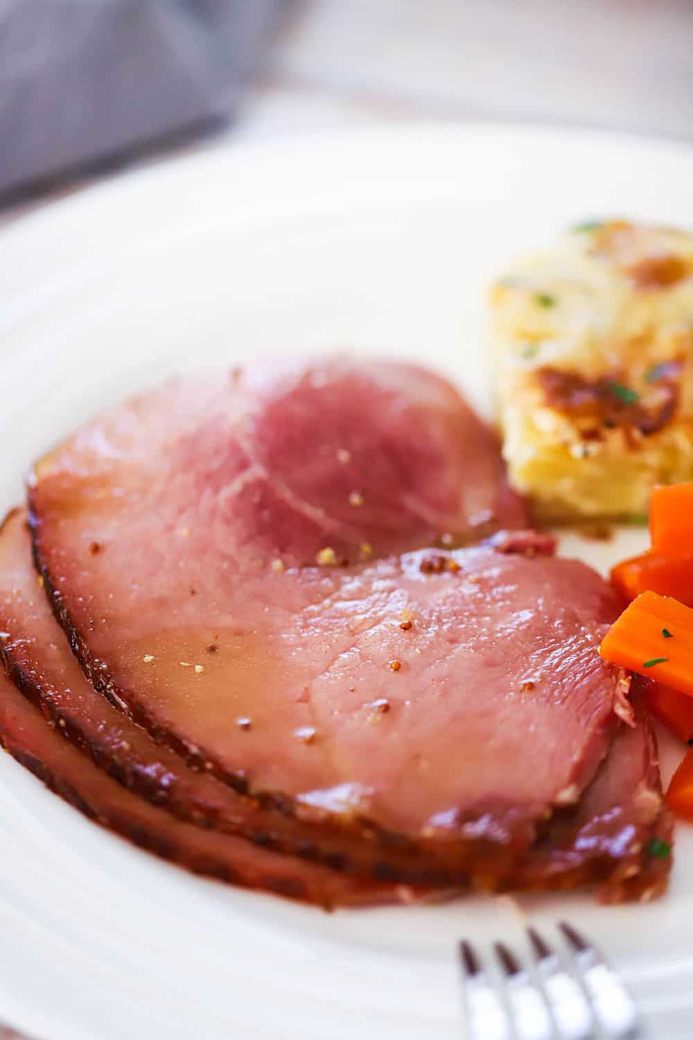 A white dinner plate filled with several slices of baked ham next to au gratin potatoes and braised carrots. 