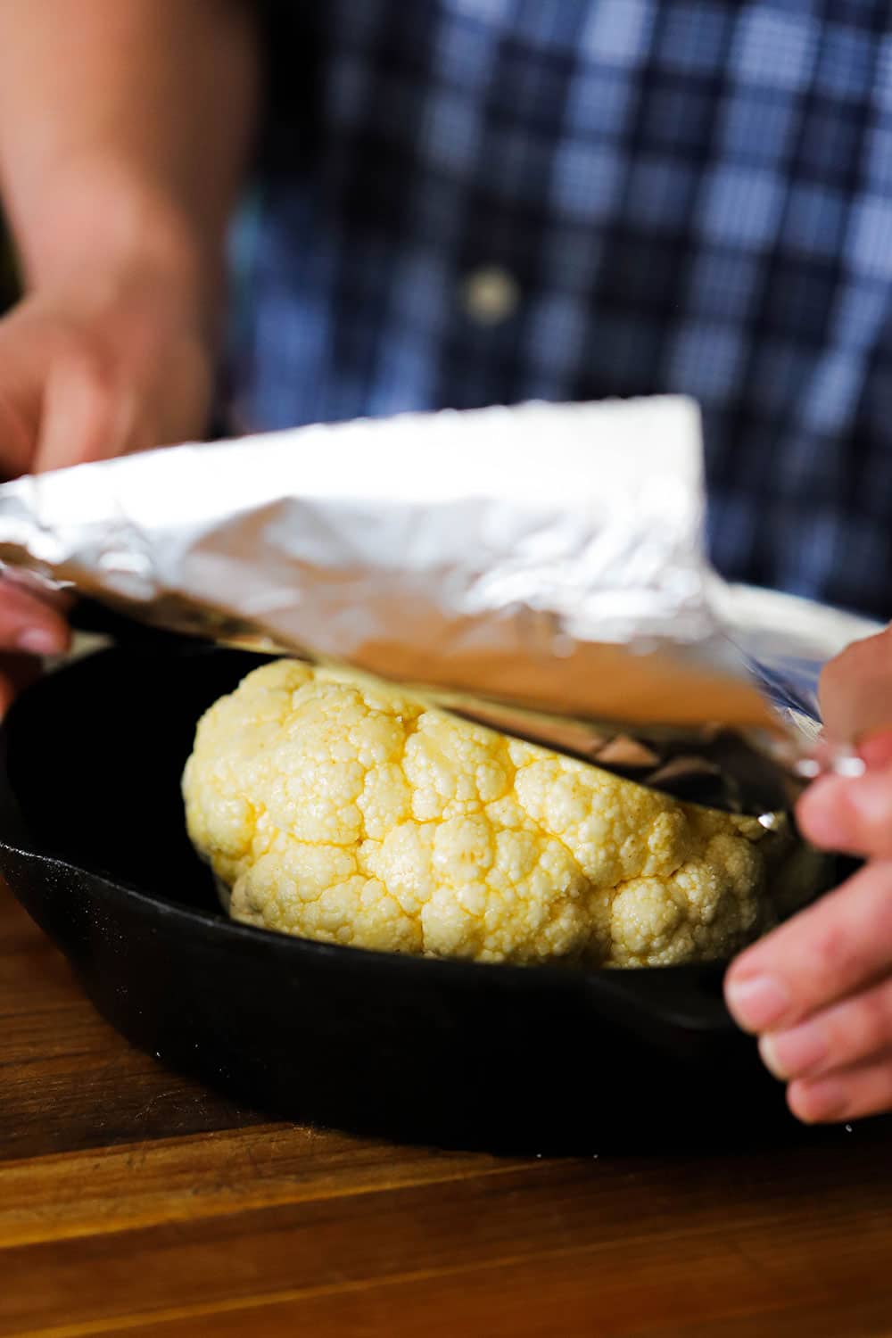 A person placing a piece of foil over a head of cauliflower that is sitting in a cast-iron skillet.