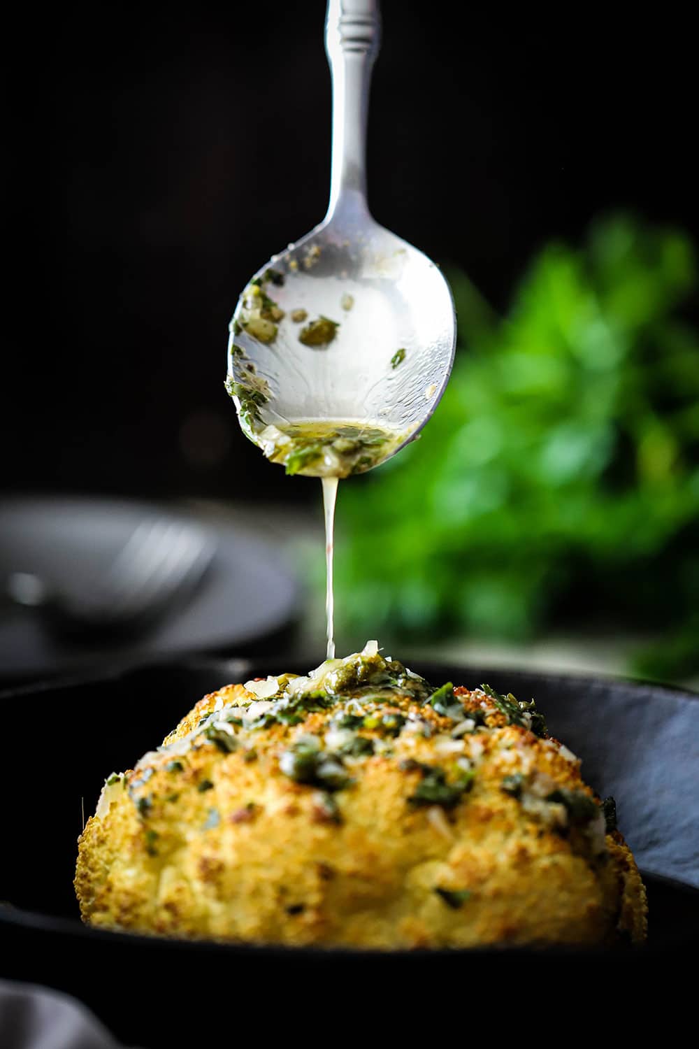 A silver serving spoon pouring a white wine butter and herb sauce over a roasted whole head of cauliflower.