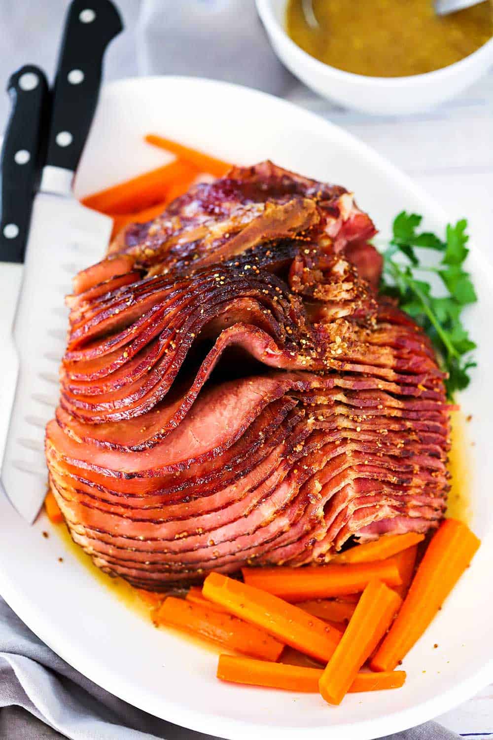 A spiral baked ham sitting on a large oval white platter with cooked carrots surrounding the roast. 