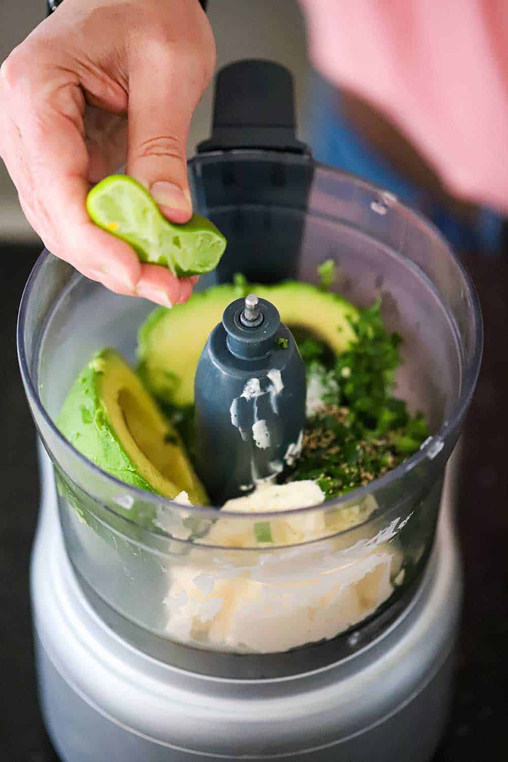 A person squeezing a cut lime into a food processor filled with avocado, mayonnaise, and cilantro. 