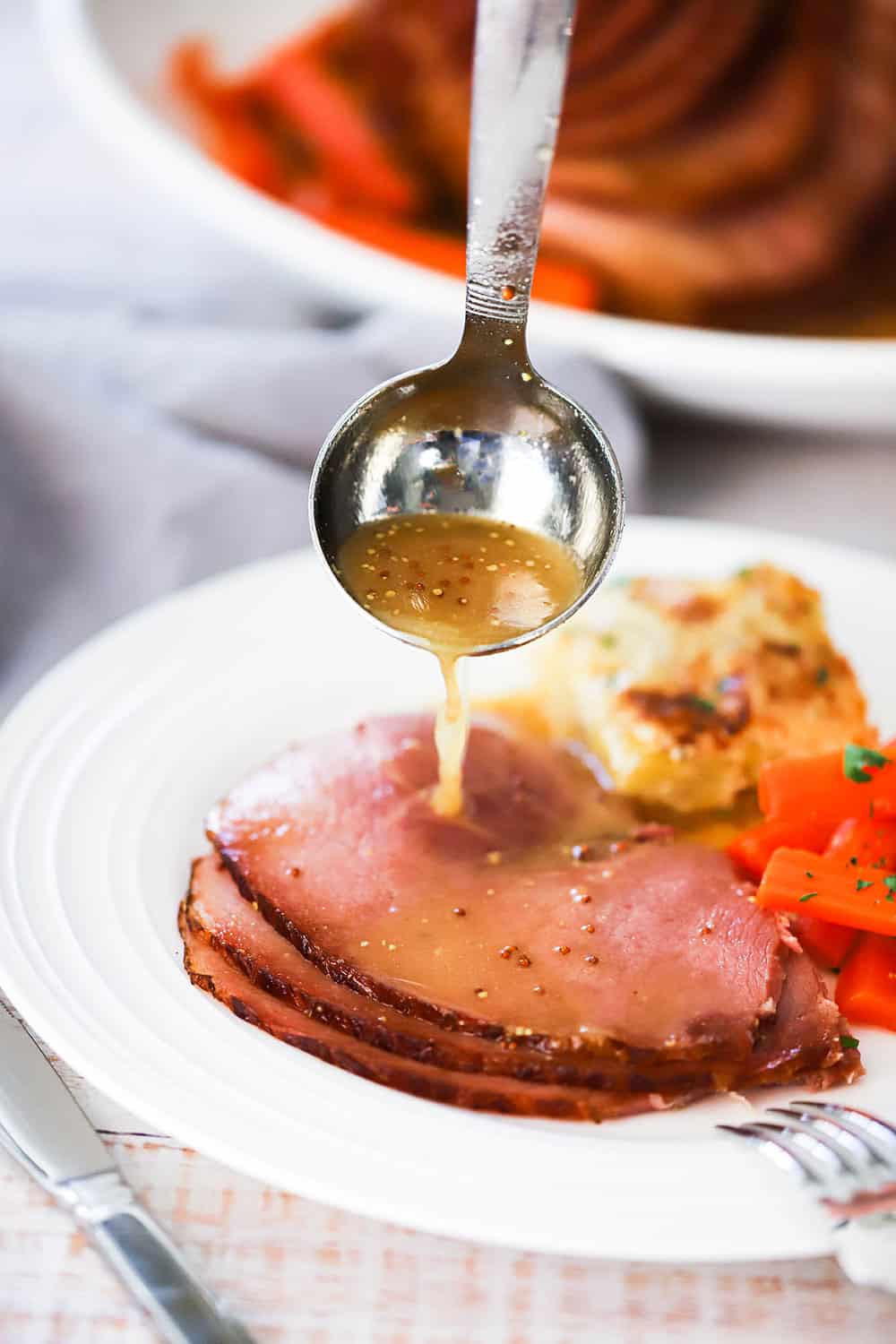 A mustard-maple sauce being poured from a small ladle onto a plate filled with several slices of baked ham. 