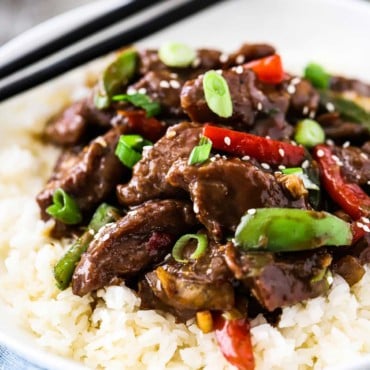 A white circular bowl filled with white rice topped with Mongolian Beef next to a pair of chop sticks.