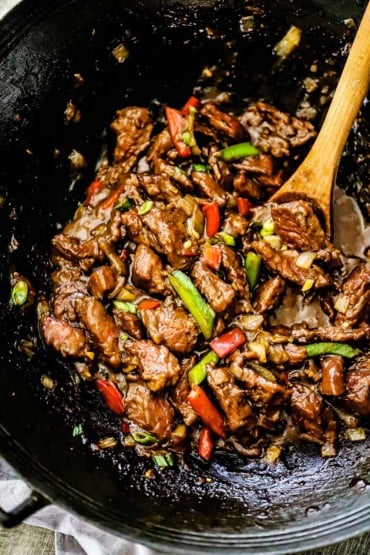 A wok filled with fully cooked mongolian beef with a wooden spoon inserted into the mixture.