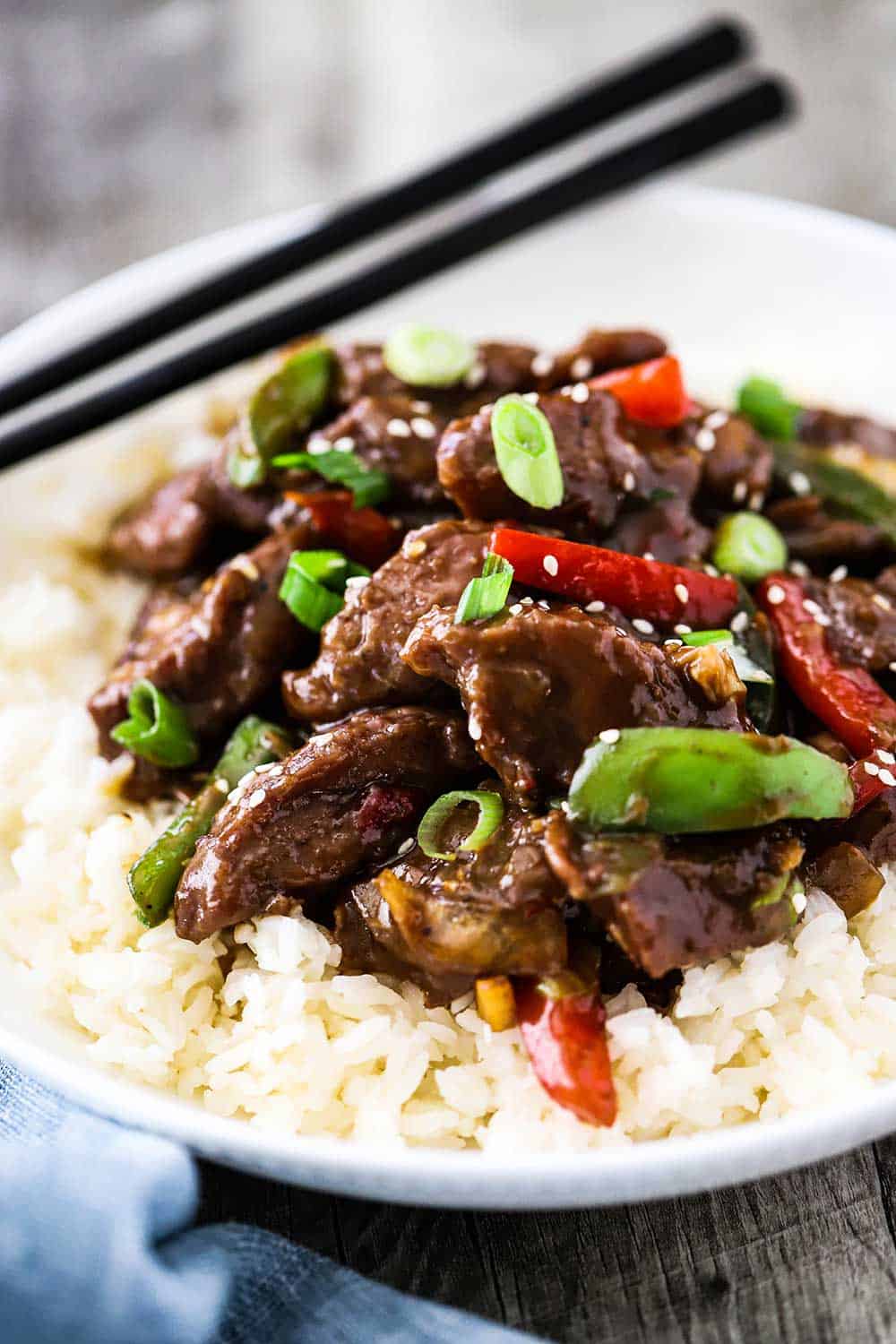 A white circular bowl filled with white rice topped with Mongolian Beef next to a pair of chop sticks. 