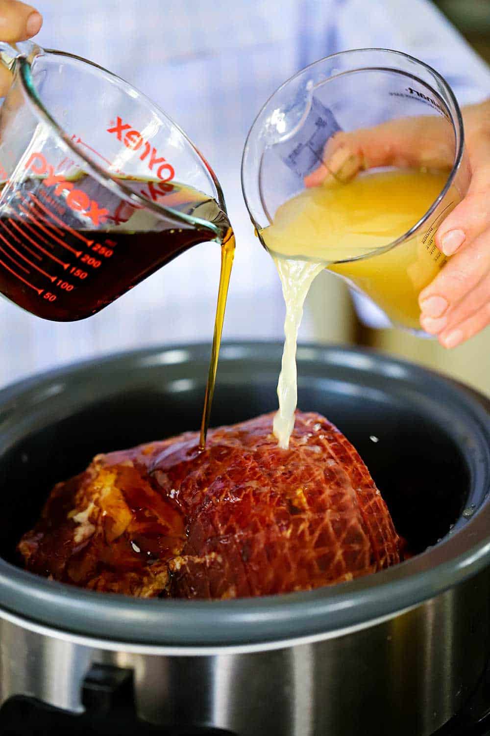 A person pouring maple syrup from a measuring cup as well as a cup of apple cider into a slow-cooker filled with a spiral smoked ham. 