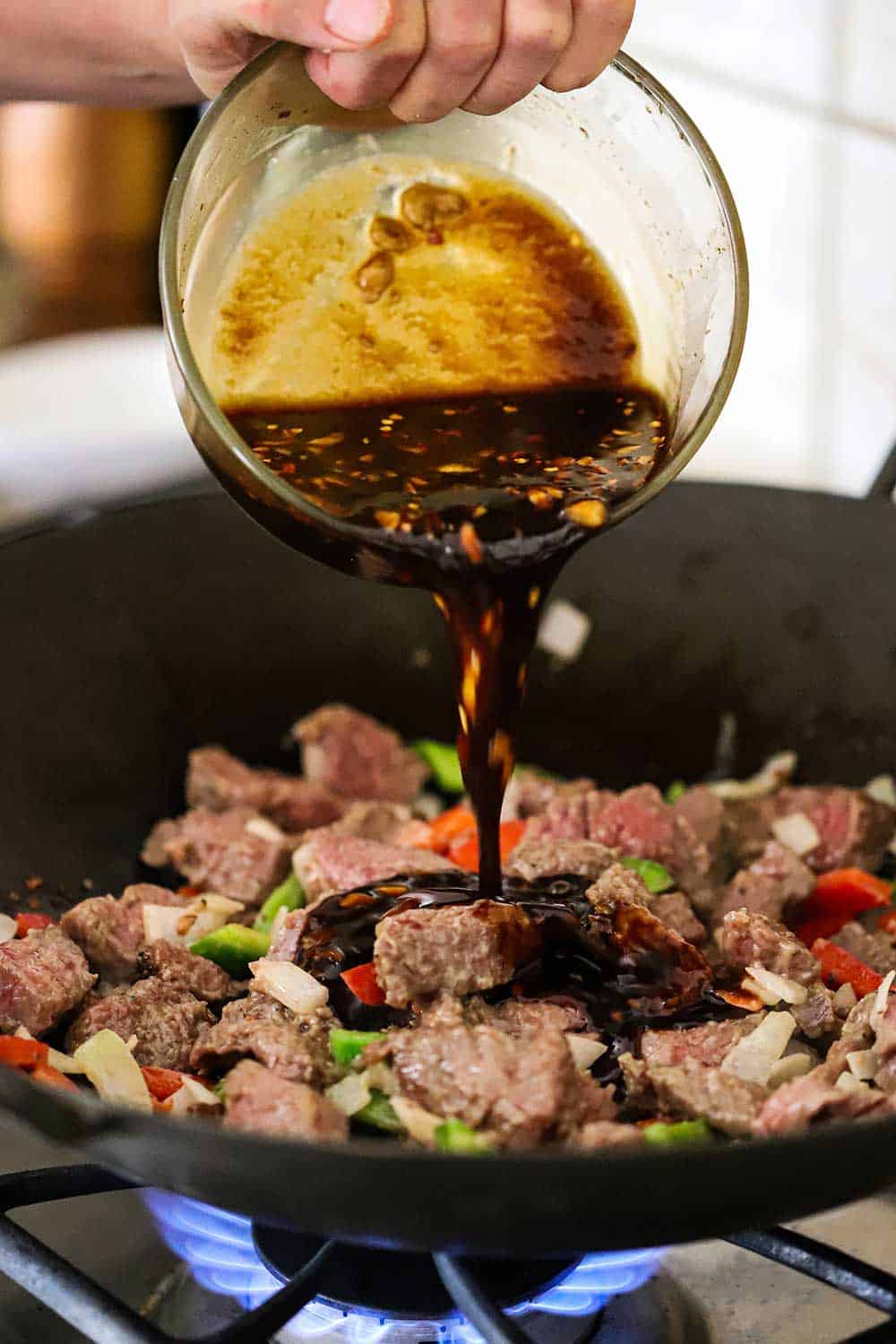 Kris is transferring the sauce from a small glass bowl into the wok filled with cooked beef and vegetables. 