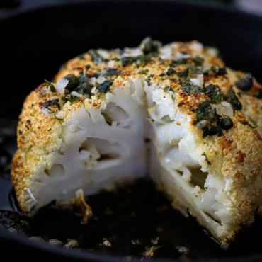 A whole head of roasted cauliflower with a slice taken out of it with a wine butter and herb sauce all over the top.