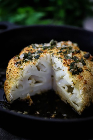 A whole head of roasted cauliflower with a slice taken out of it with a wine butter and herb sauce all over the top.