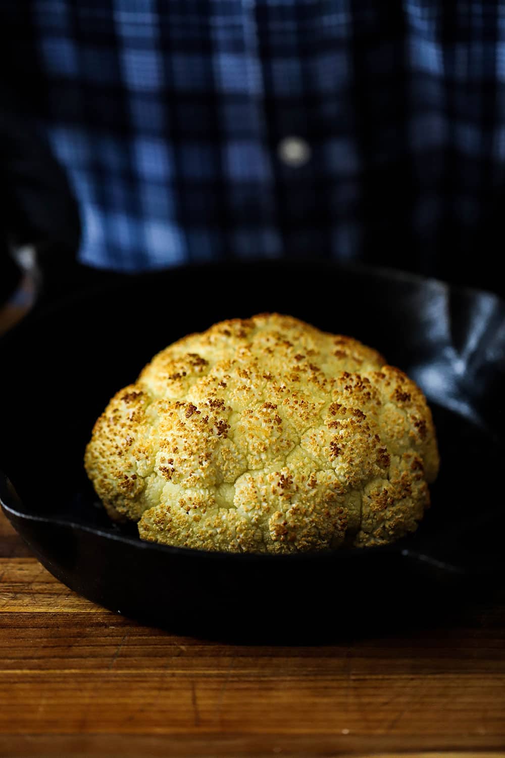 A roasted whole head of cauliflower in a cast-iron skillet being held by a person in a blue shirt.