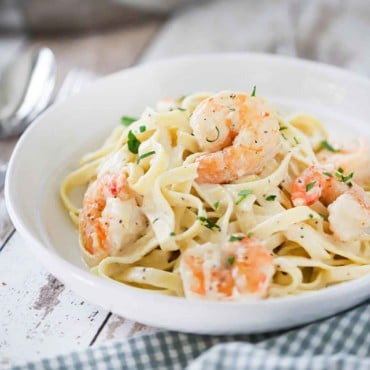 A white individual pasta bowl filled with shrimp fettuccine alfredo next to a large silver skillet filled with the same.