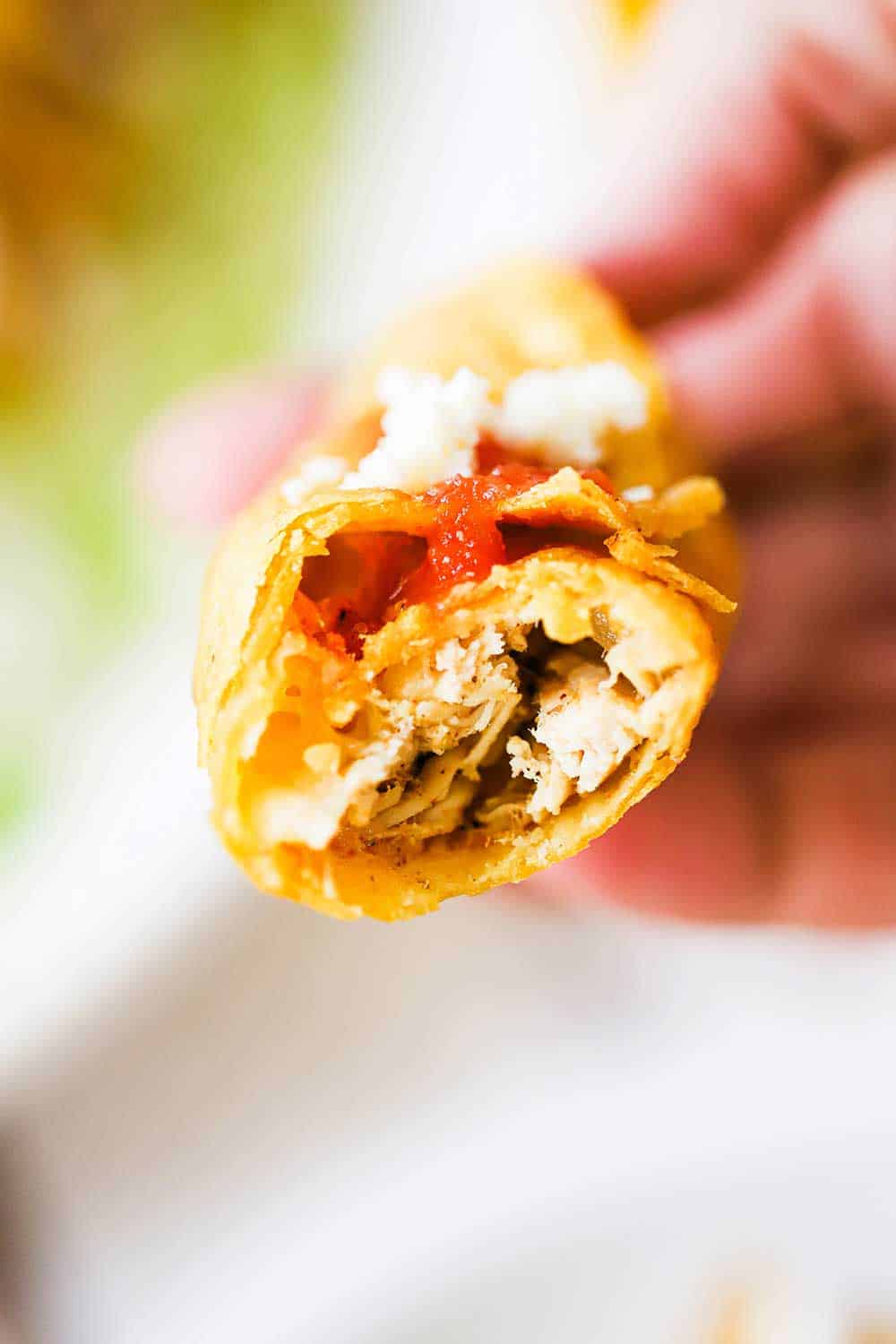 A hand holding up a deep-fried chicken flauta with a bite taken out of it so the chicken filling is visible. 