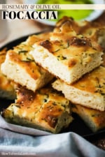 A black plate with a stack of square pieces of focaccia bread with rosemary and olive oil on it.