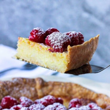 As slice of Chess Pie topped with raspberries and a sprinkling of powdered sugar being raised from the pie on a metal spatula.