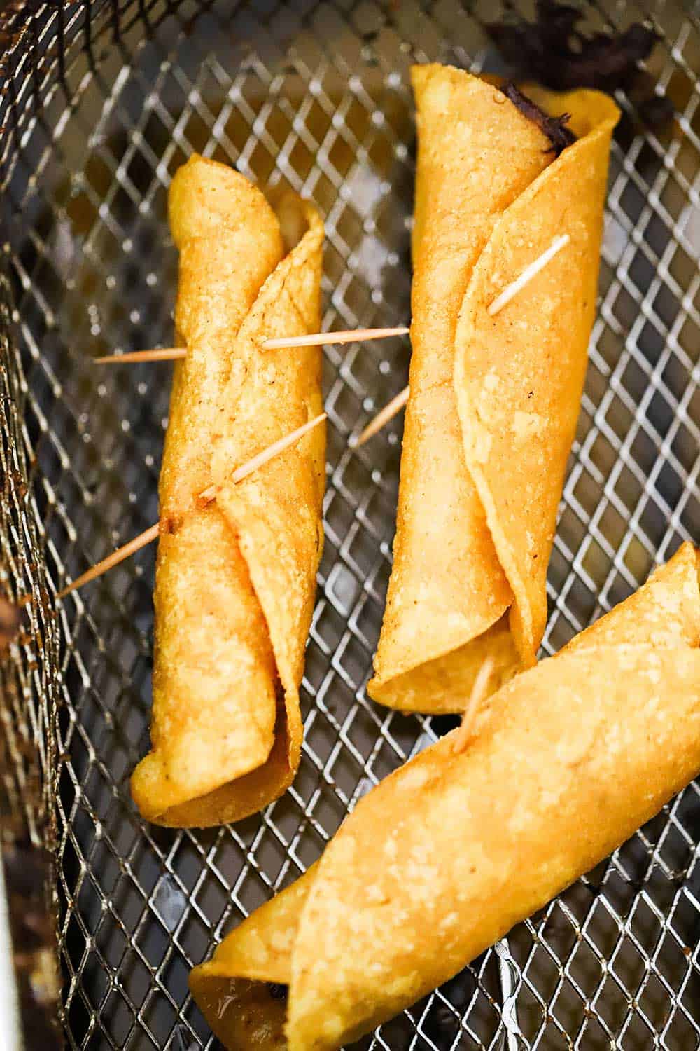 Three rolled fried flautas in a deep fryer basket. 