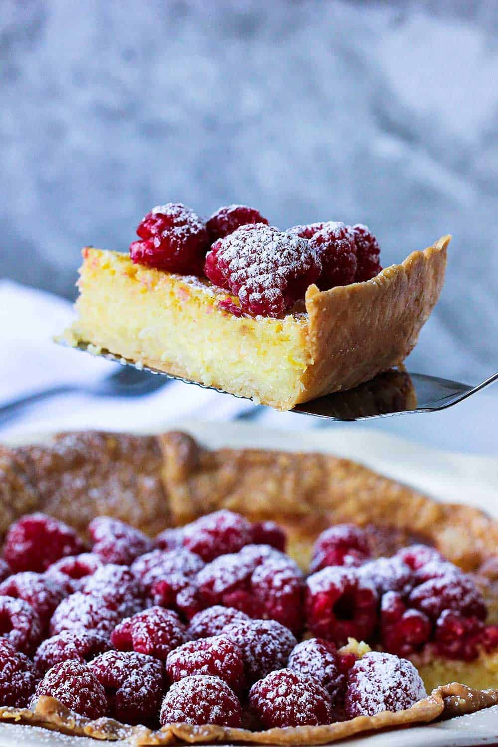 As slice of Chess Pie topped with raspberries and a sprinkling of powdered sugar being raised from the pie on a metal spatula.