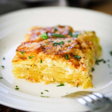 A square serving of potatoes dauphinoise on a white plate garnished with chopped parsley.
