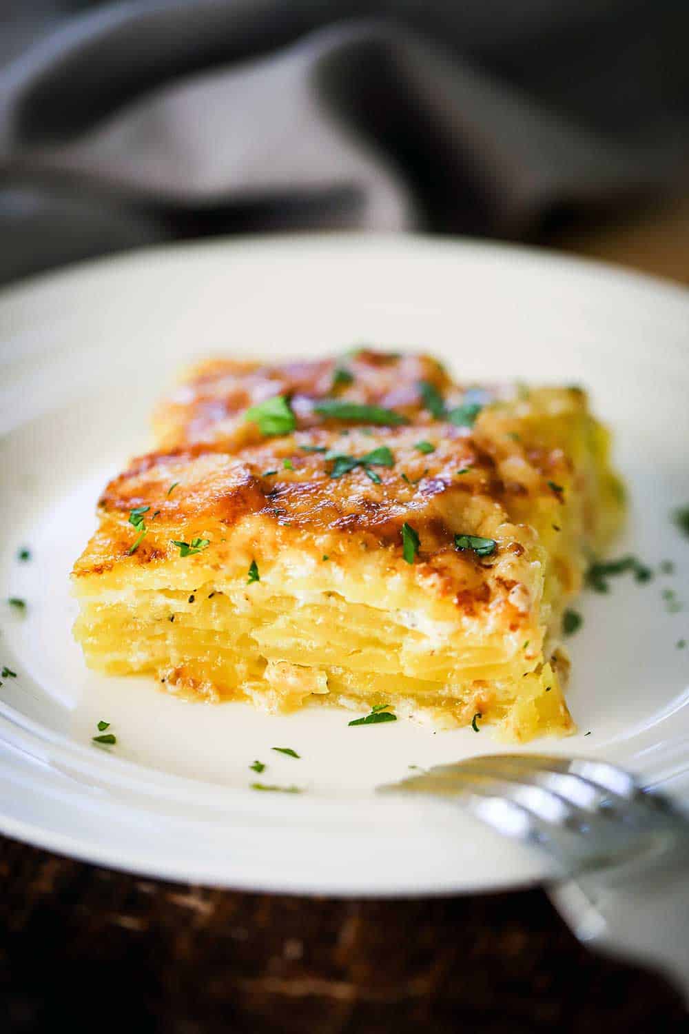 A square serving of potatoes dauphinoise on a white plate garnished with chopped parsley.