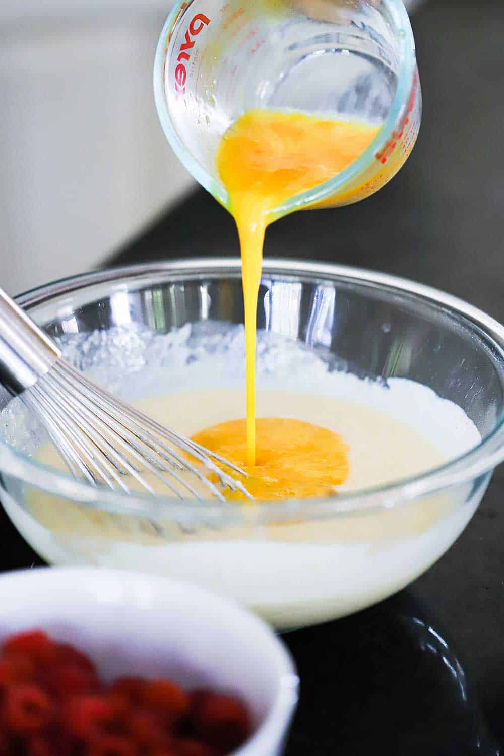 Beaten eggs being poured from a measuring cup into a bowl of white colored batter.