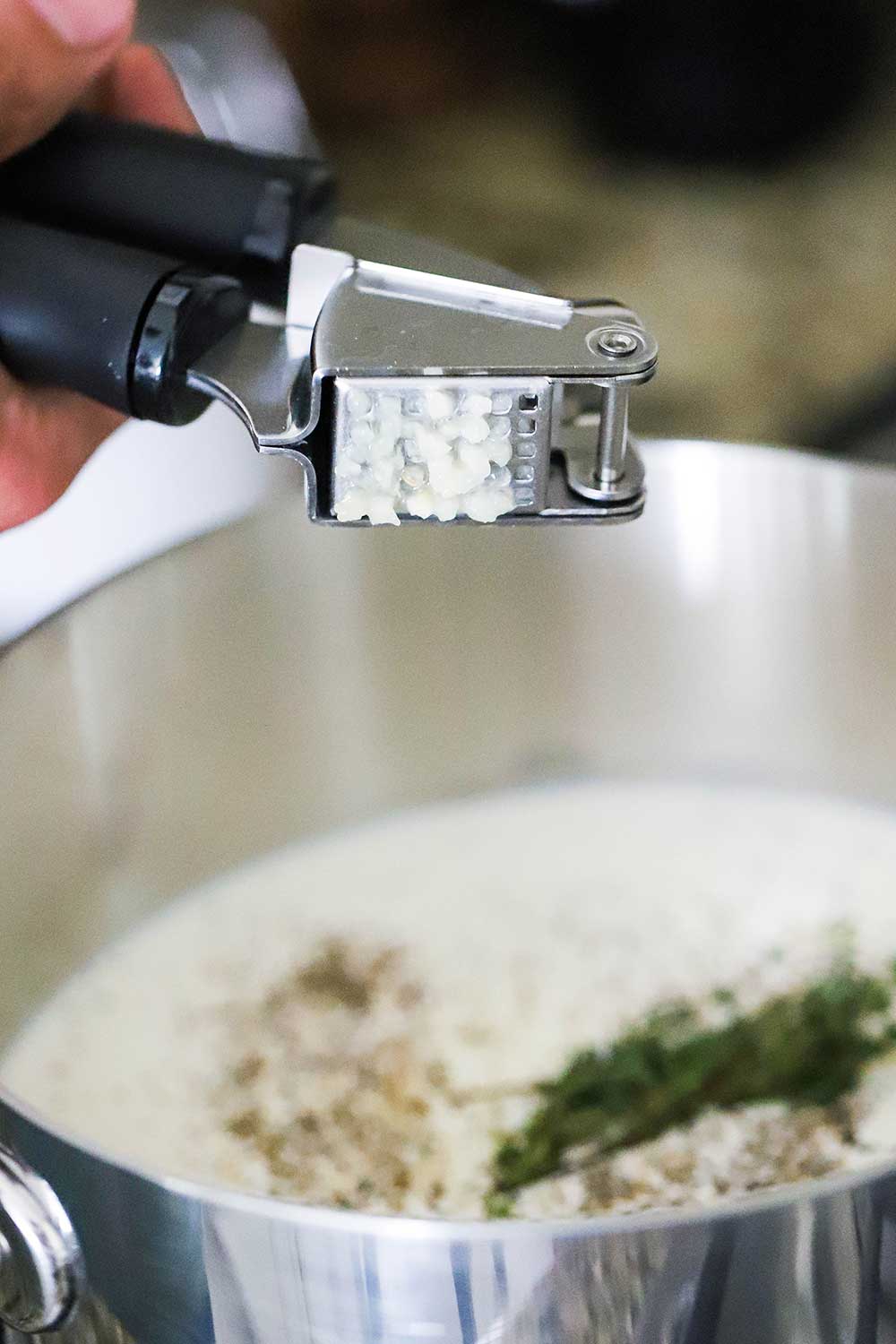 A person squeezing garlic out of a garlic press into a pan filled with cream and herbs.