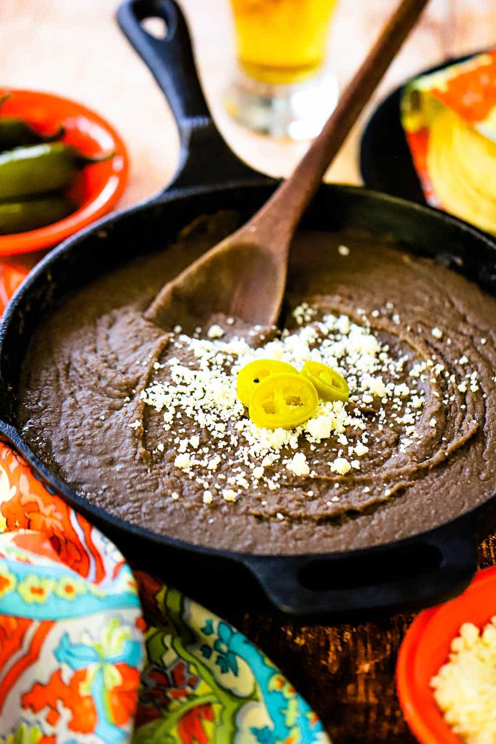 A cast-iron skillet filled with refried beans and topped with crumbled Cojita cheese and several jalapeno slices.